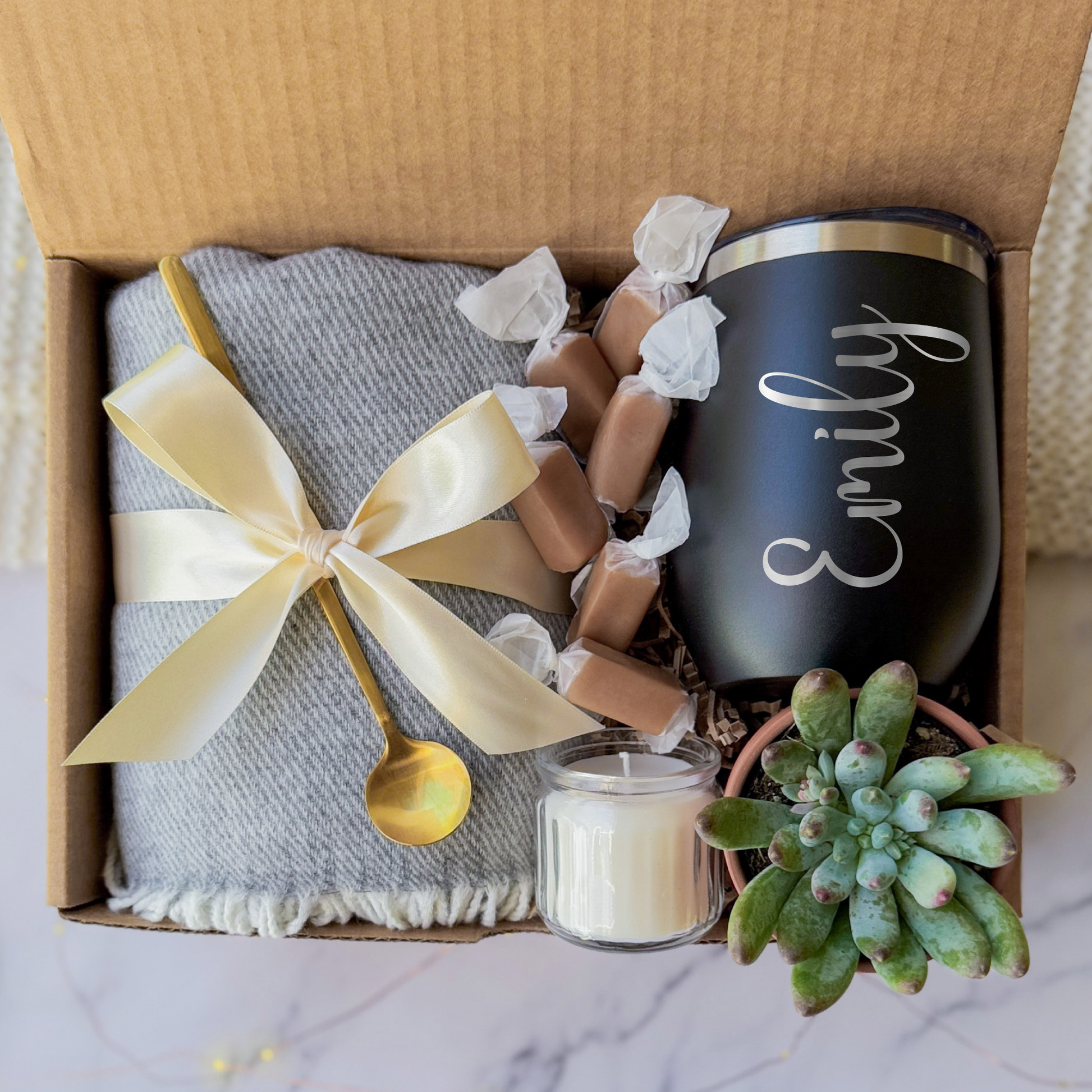 Gift box containing a personalized black tumbler with the name engraved, a soft gray blanket tied with a cream ribbon, a small succulent plant, a golden spoon, a scented candle, and caramel toffees, arranged neatly in a cardboard box.