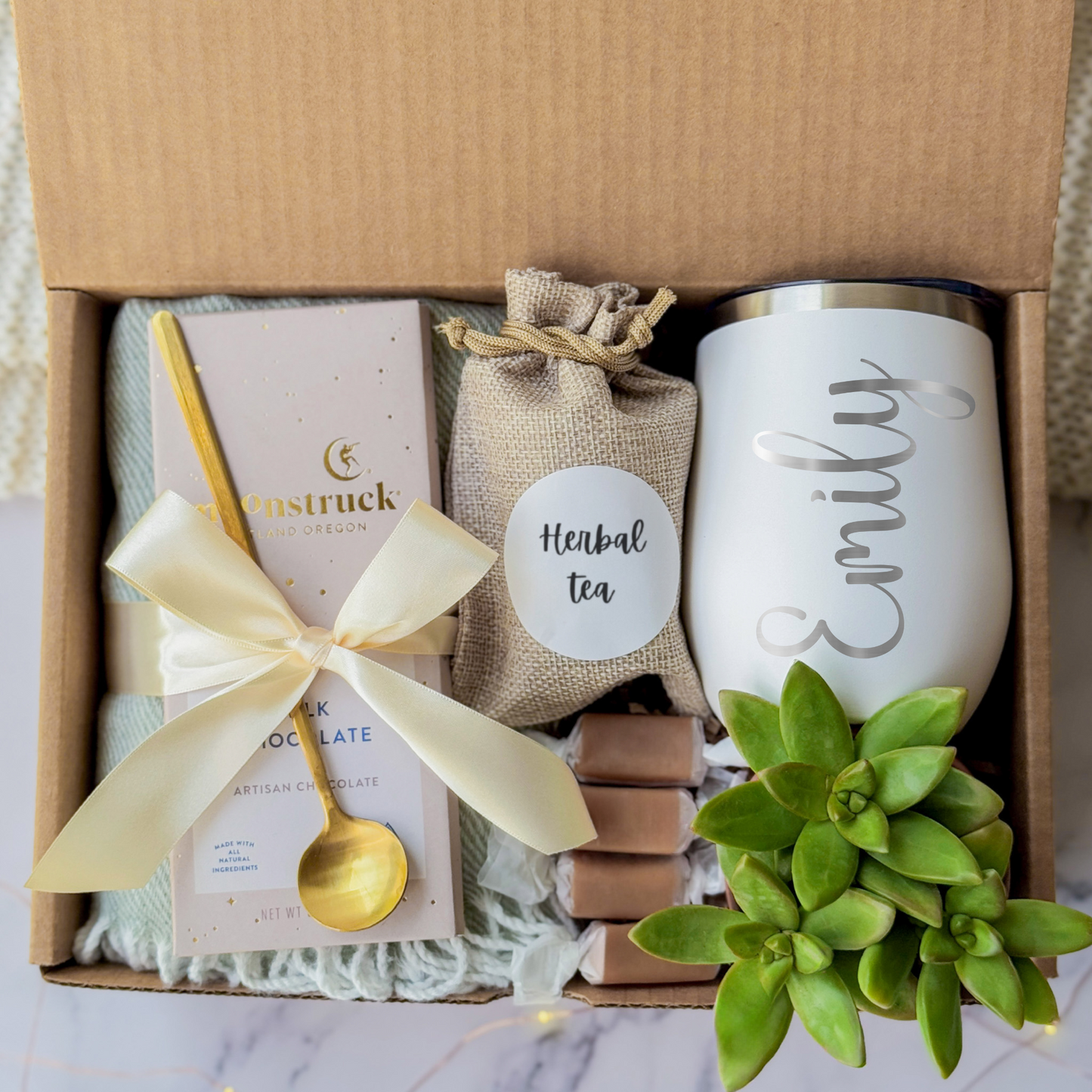 A gift box containing a personalized white tumbler with name engraving written in cursive, a green succulent, a pouch labeled "Herbal Tea," a milk chocolate bar tied with a gold spoon and a cream ribbon, and caramel candies. The items are arranged on a soft blanket inside a brown box, presented on a marble surface.