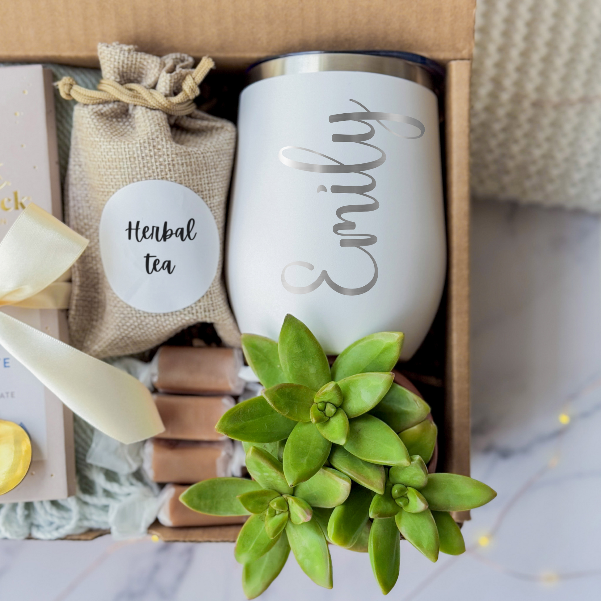 A gift box containing a personalized white tumbler with name engraving written in cursive, a green succulent, a pouch labeled "Herbal Tea," a milk chocolate bar tied with a gold spoon and a cream ribbon, and caramel candies. The items are arranged on a soft blanket inside a brown box, presented on a marble surface.