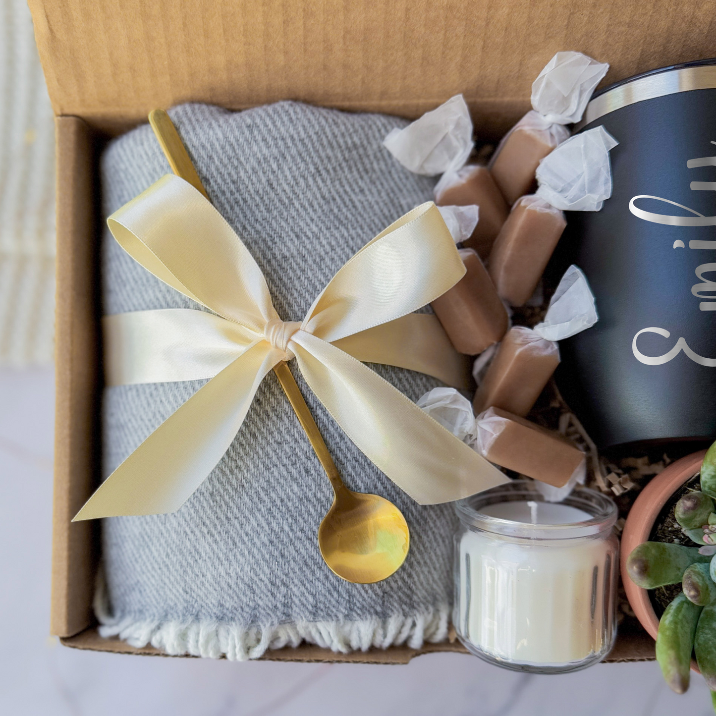 Gift box containing a personalized black tumbler with the name engraved, a soft gray blanket tied with a cream ribbon, a small succulent plant, a golden spoon, a scented candle, and caramel toffees, arranged neatly in a cardboard box.