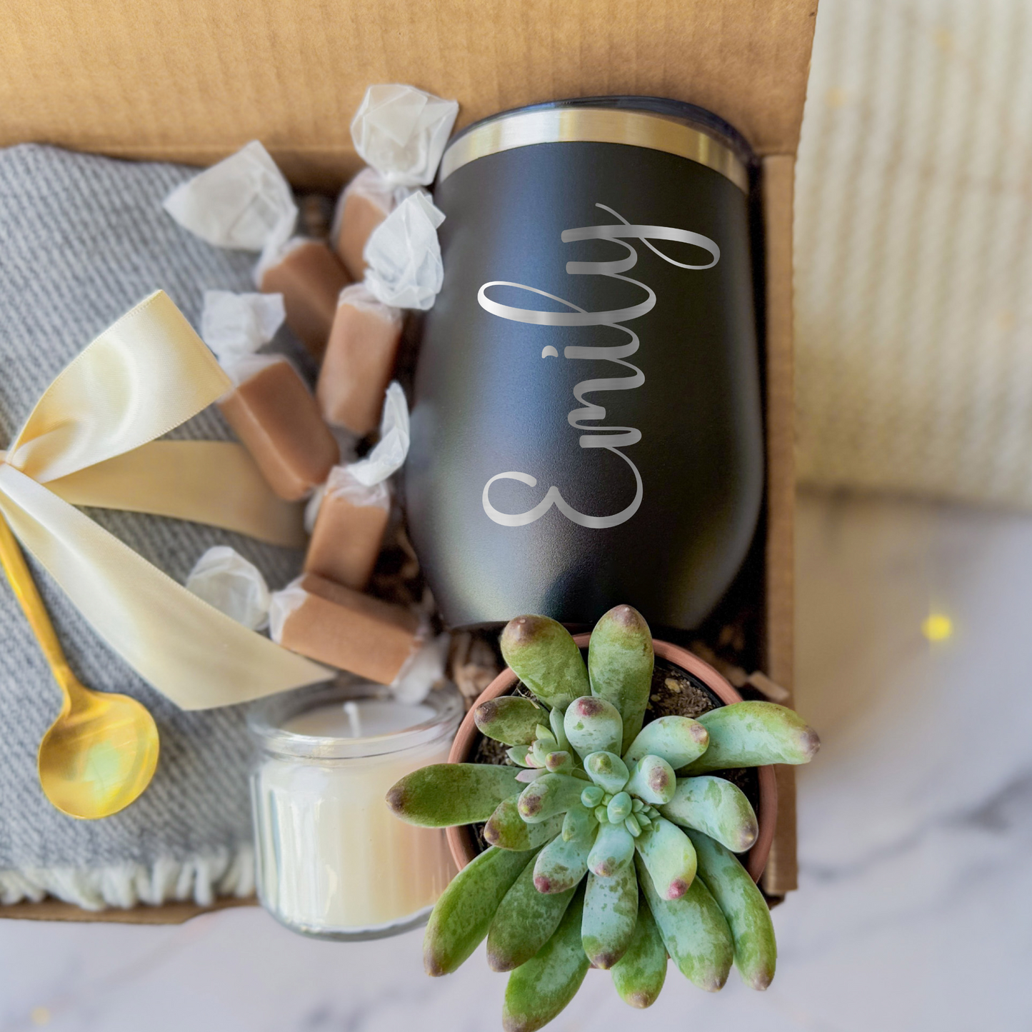 Gift box containing a personalized black tumbler with the name engraved, a soft gray blanket tied with a cream ribbon, a small succulent plant, a golden spoon, a scented candle, and caramel toffees, arranged neatly in a cardboard box.