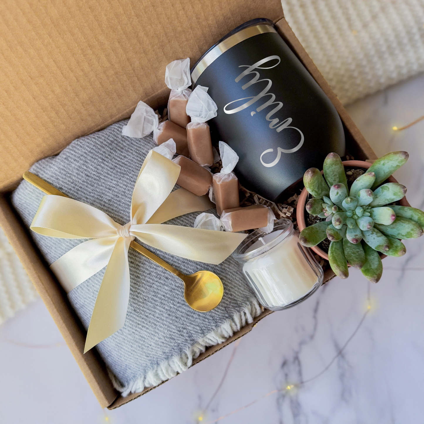 Gift box containing a personalized black tumbler with the name engraved, a soft gray blanket tied with a cream ribbon, a small succulent plant, a golden spoon, a scented candle, and caramel toffees, arranged neatly in a cardboard box.
