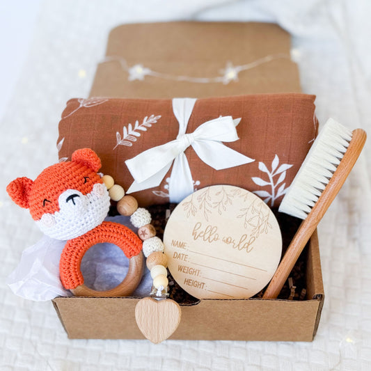 A beautifully arranged baby gift box featuring a rust-colored swaddle with a white leaf print tied with a ribbon, a wooden baby brush, a crocheted orange fox rattle, a wooden teether with beads and a heart detail, and a round wooden "Hello World" milestone card for recording baby’s name, date, weight, and height. The items are placed neatly inside a brown box on a soft white fabric background.