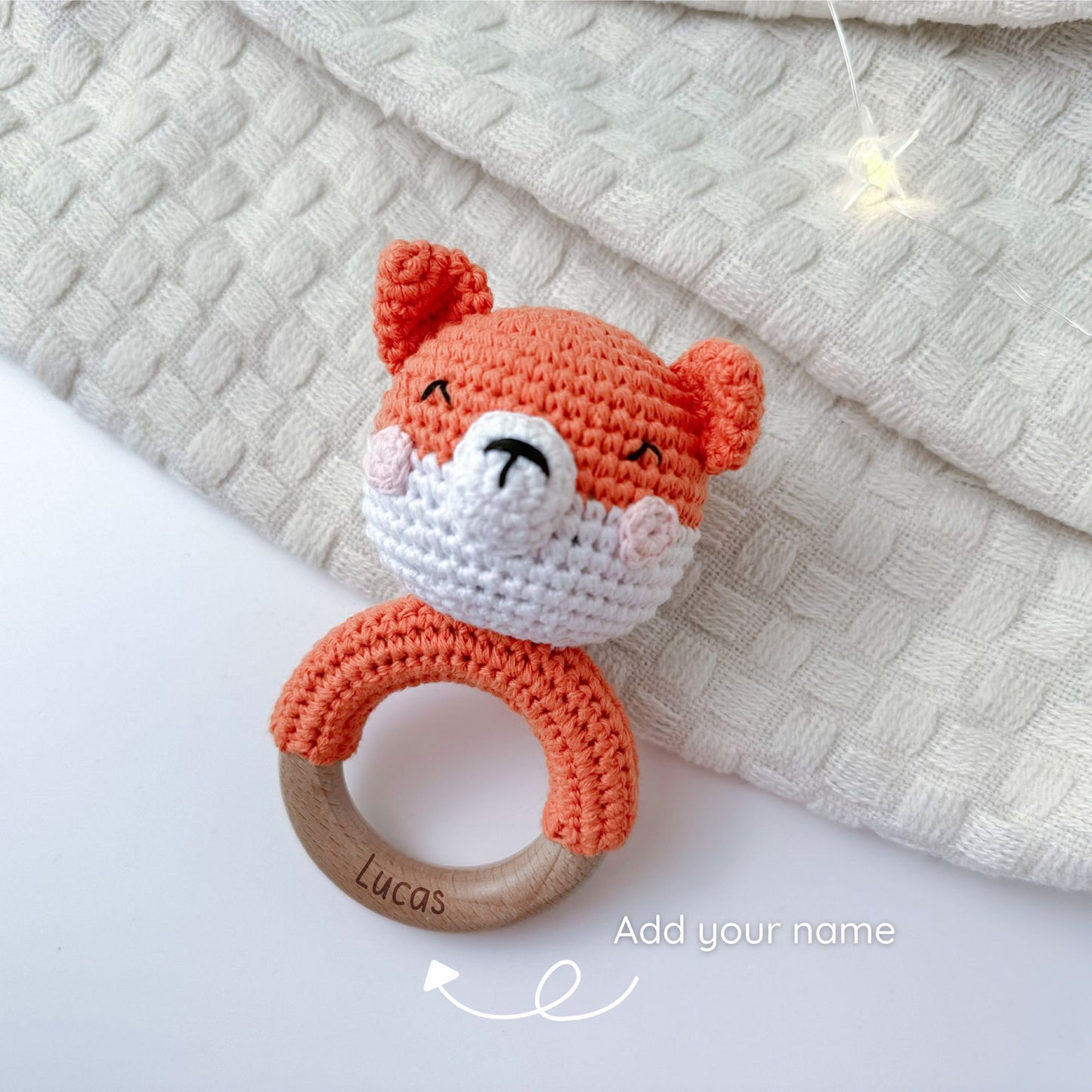 Crocheted orange fox baby rattle with a wooden ring, personalized with the name 'Lucas' engraved on the ring, placed on a soft, textured beige fabric background.