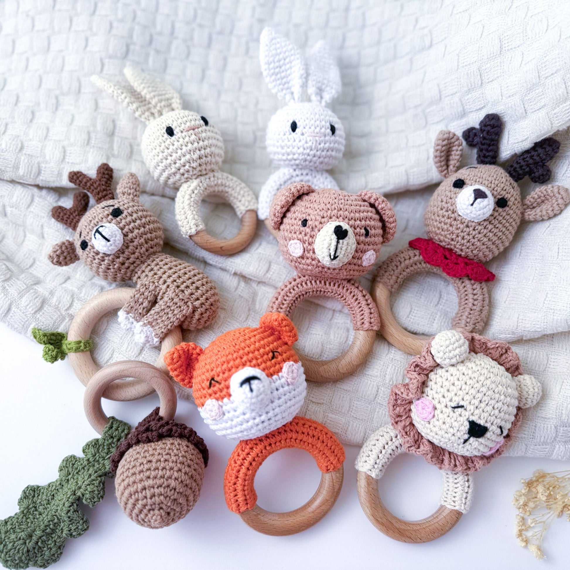 Collection of crocheted baby rattles featuring animal designs, including a fox, bear, lion, deer, and rabbits, along with a crocheted acorn and leaf, all with wooden rings and placed on a soft beige fabric background.