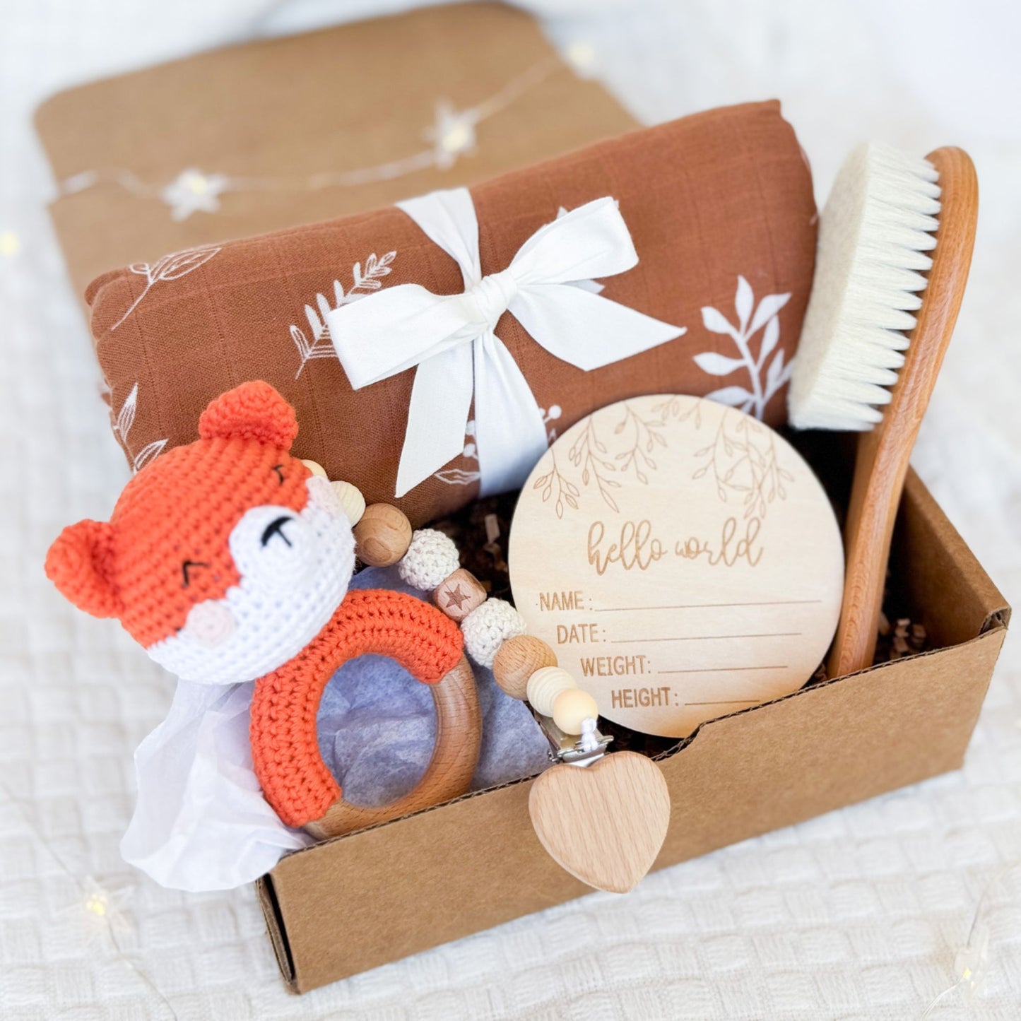 A beautifully arranged baby gift box featuring a rust-colored swaddle with a white leaf print tied with a ribbon, a wooden baby brush, a crocheted orange fox rattle, a wooden teether with beads and a heart detail, and a round wooden "Hello World" milestone card for recording baby’s name, date, weight, and height. The items are placed neatly inside a brown box on a soft white fabric background.