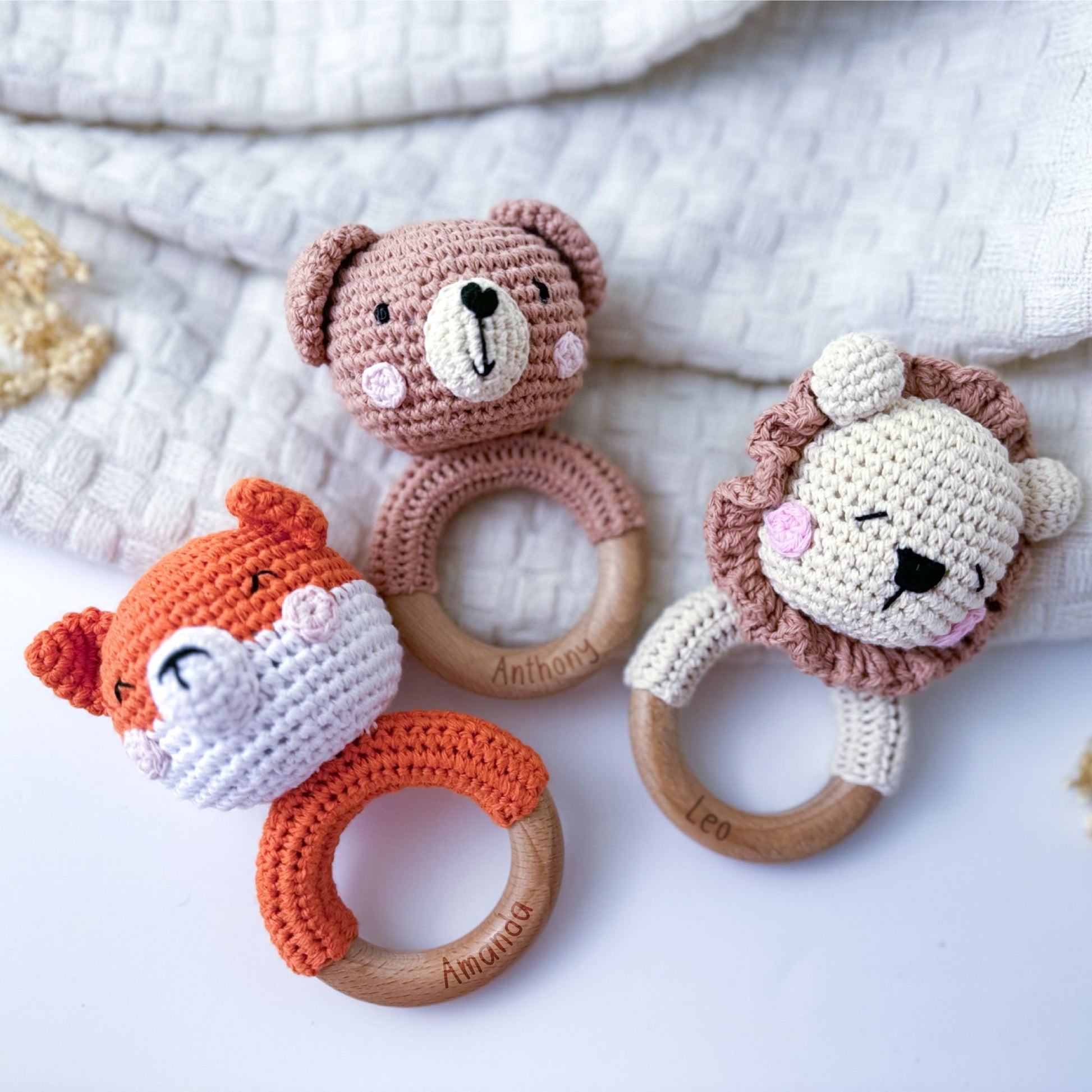 Set of three crocheted baby rattles featuring animal designs: an orange fox, a beige bear, and a light brown lion, each with a personalized wooden ring engraved with names (Anthony, Leo, and Annabella), displayed on a soft, textured beige fabric background.