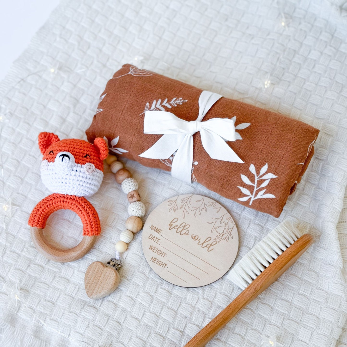 A neatly arranged baby gift set displayed on a textured white background. The set includes a rust-colored swaddle with white leaf prints tied with a white ribbon, a crocheted orange fox rattle with a wooden ring, a wooden teether with beads and a heart-shaped detail, a round wooden "Hello World" milestone card for recording baby details, and a wooden baby brush with soft bristles. The items are thoughtfully laid out to showcase their quality and charm.